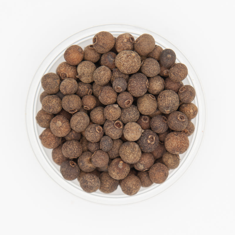 Clear glass bowl filled with brown allspice berries on a white background